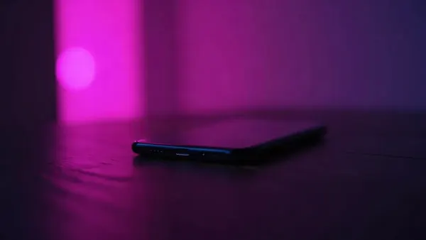 Smartphone na tmavém dřevěném stole, obklopený fialovými a růžovými odlesky (ilustrace k článku)